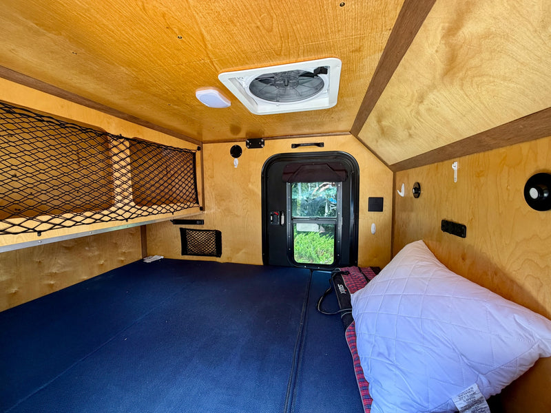 Compact RV rental interior with wooden walls, blue bed, ceiling fan, and storage nets, ready for your next adventure. — photo 9
