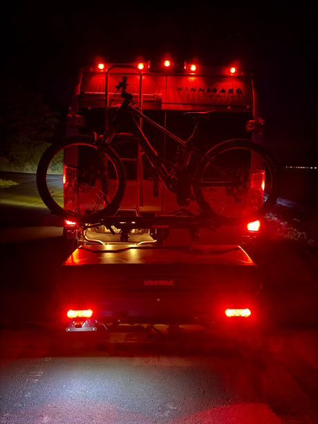 An RV rental at night, with a mountain bike on a Yakima rack and cargo box attached, illuminated by bright red taillights.