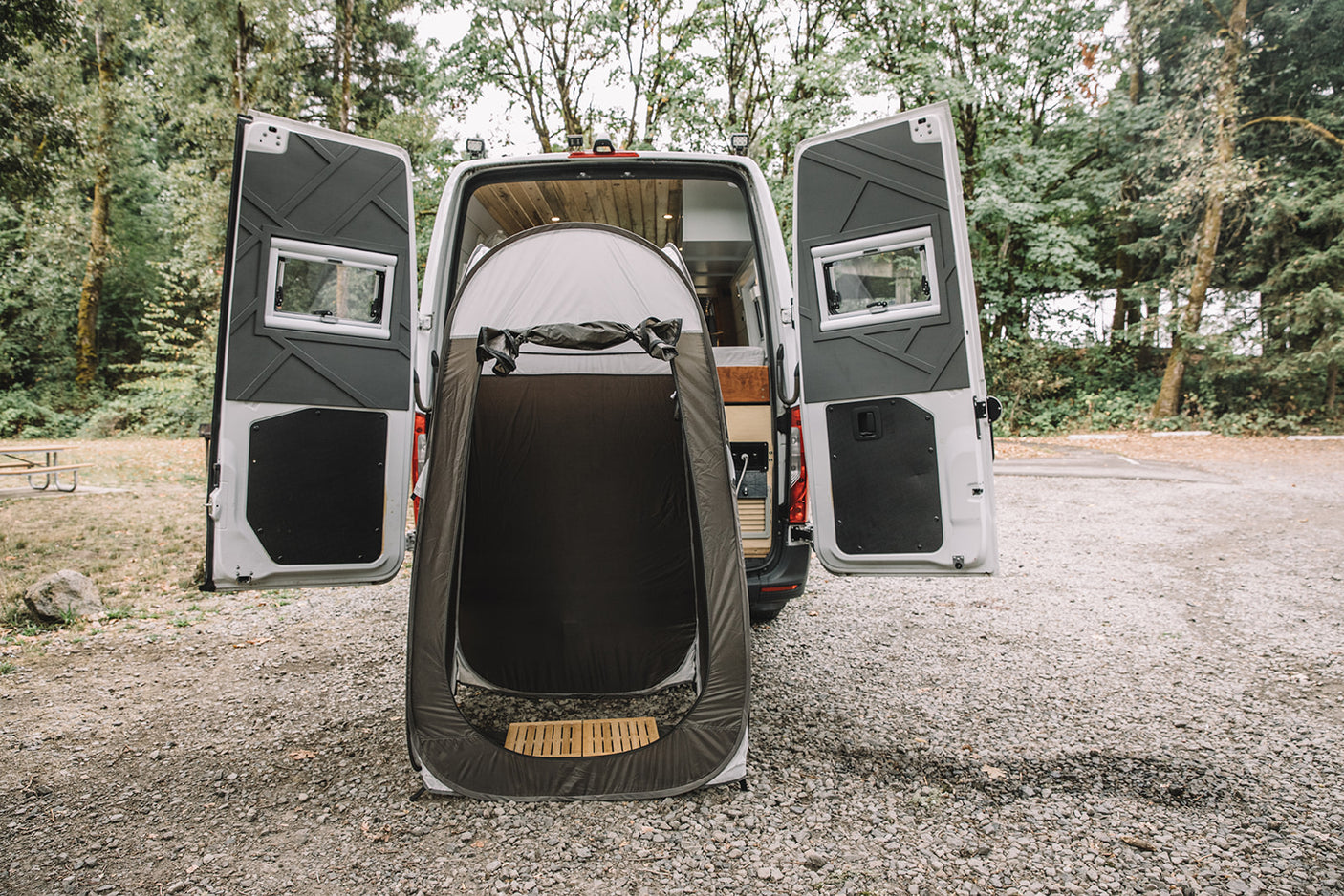An RV rental's rear opens to a grey portable shower tent on gravel. Perfect for outdoor camping and privacy.