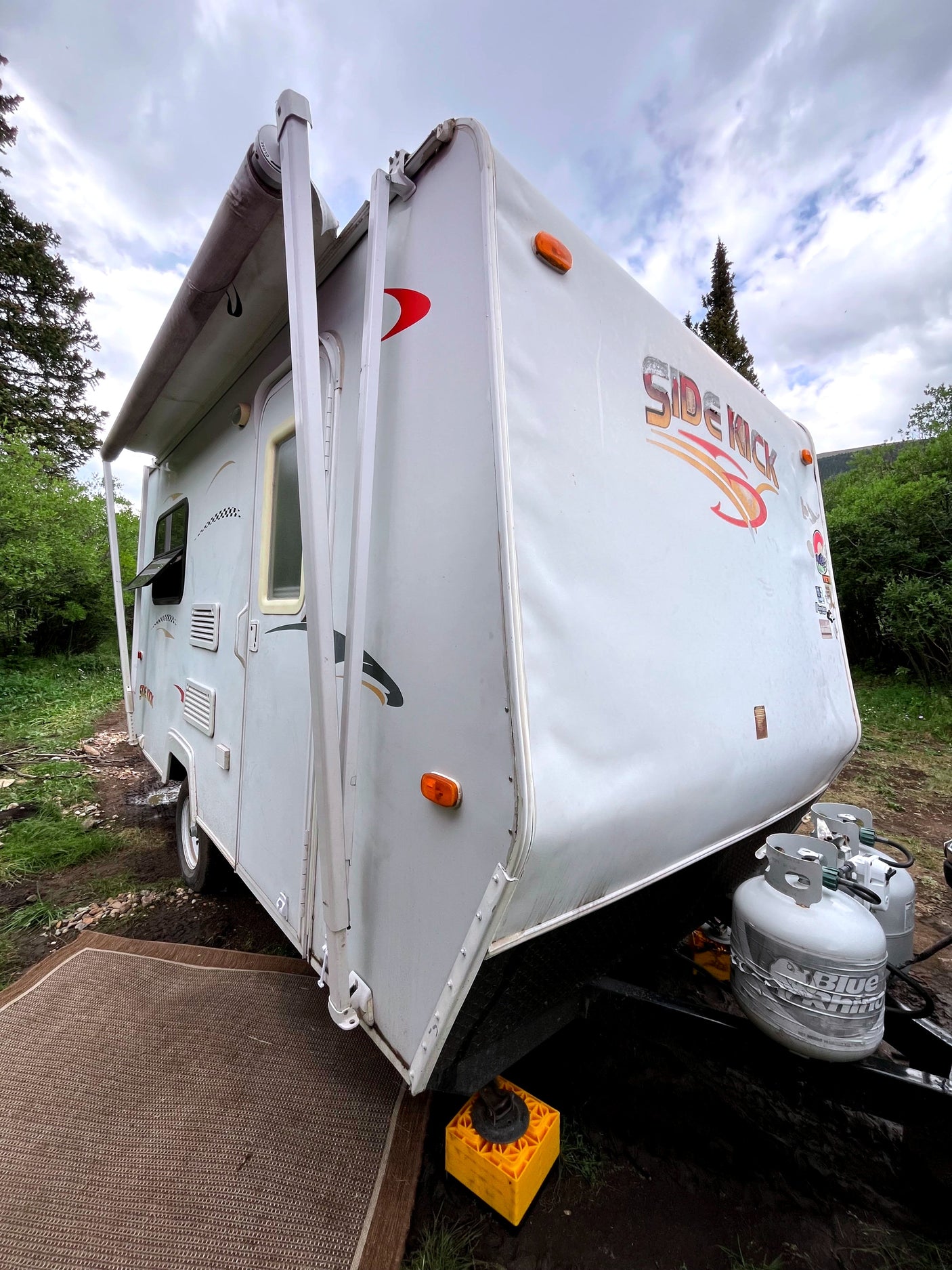 White "Side Kick" travel trailer RV rental with extended awning, propane tanks, parked in a treed, muddy setting.
