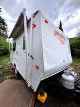 White "Side Kick" travel trailer RV rental with extended awning, propane tanks, parked in a treed, muddy setting.