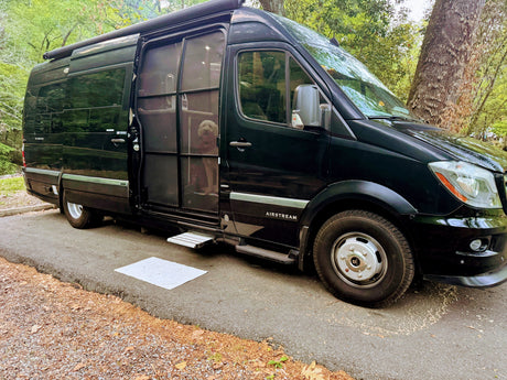 An Airstream Interstate RV rental is parked with its side door open, revealing a dog peering out from the camper van at a shady campsite.