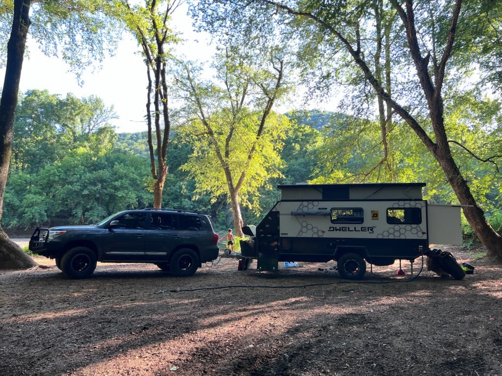 An SUV tows a pop-top off-road camper, like an rv rental, at a sunny wooded riverside campsite. Ready for adventure camping.
