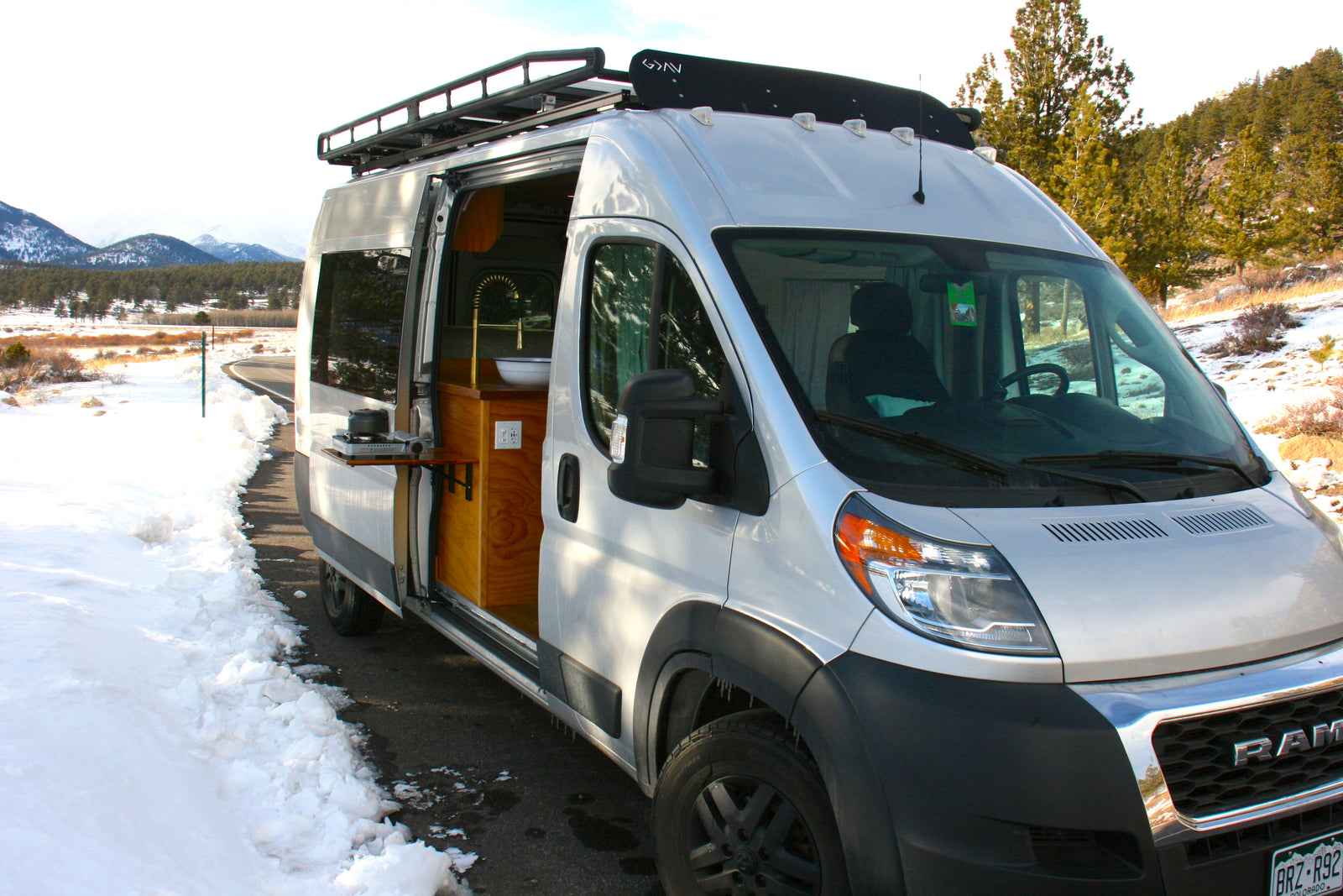 Silver RV rental van, with side door open to reveal a compact kitchen, parked by a snowy road amidst mountains.