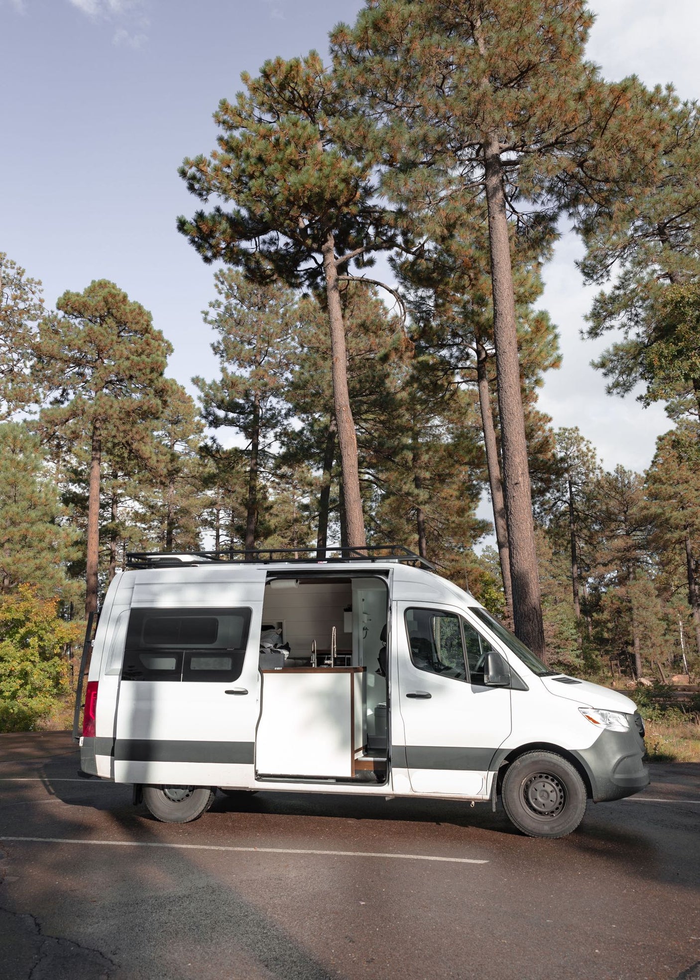 White camper van rv rental parked in a pine forest. Side door open, revealing a modern interior with kitchen setup.