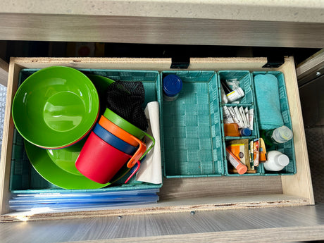 RV rental drawer featuring organized camping kitchenware: bright dishes, cups, cleaning cloths, and travel essentials in teal bins.
