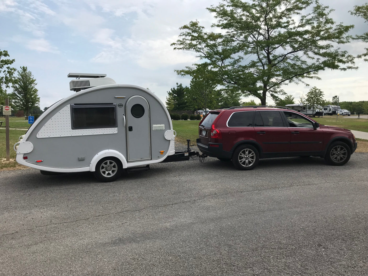 A red SUV tows a sleek grey teardrop RV rental, parked in a scenic campground ready for adventures.