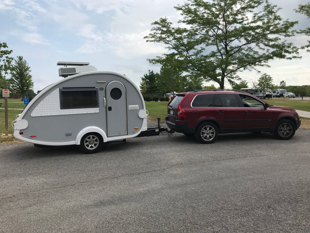 A red SUV tows a sleek grey teardrop RV rental, parked in a scenic campground ready for adventures.