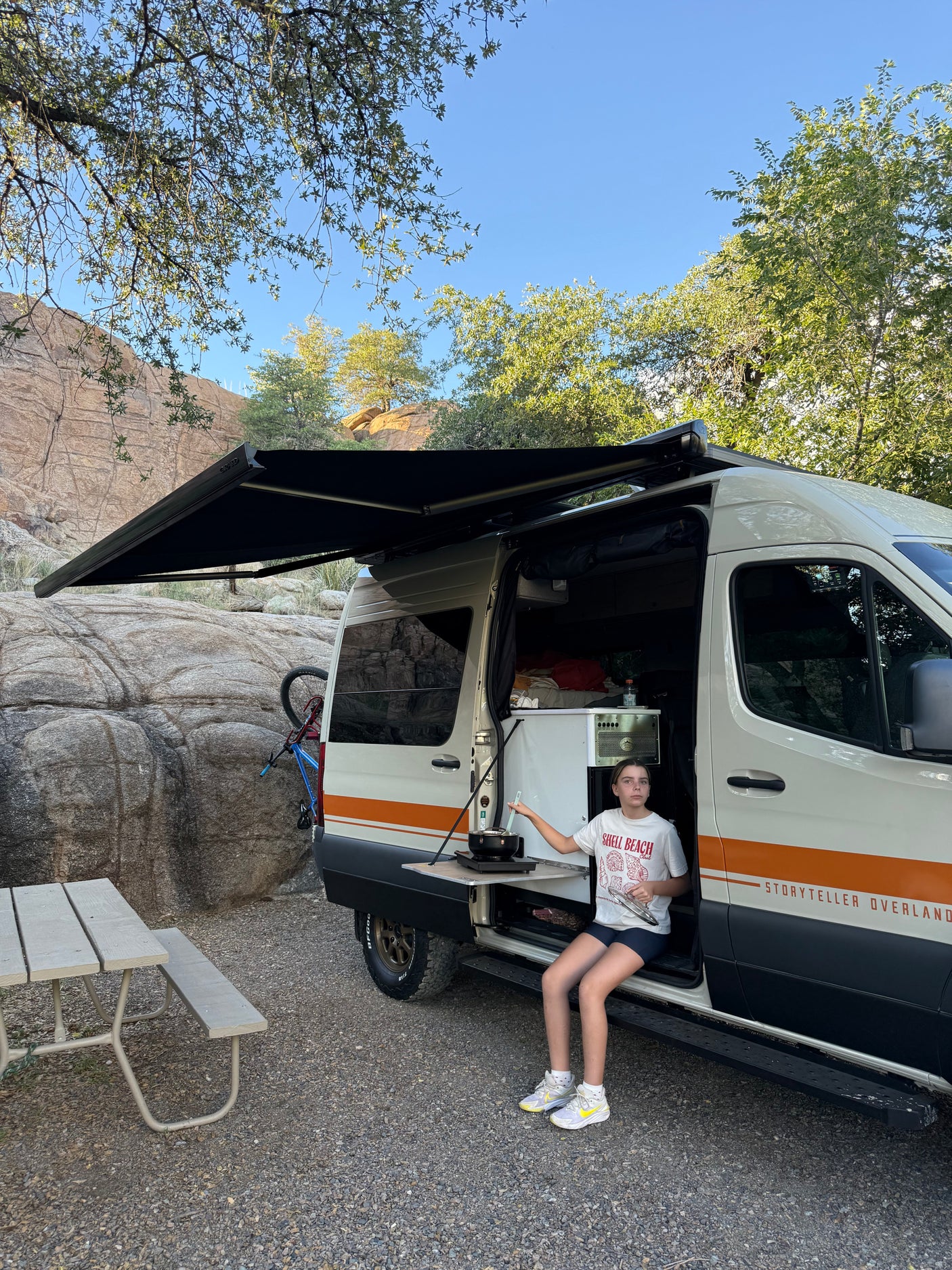 Girl cooking outside an RV rental van at a rocky campsite with an extended awning & bike.