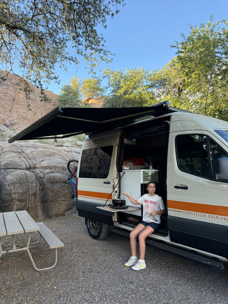 Girl cooking outside an RV rental van at a rocky campsite with an extended awning & bike.