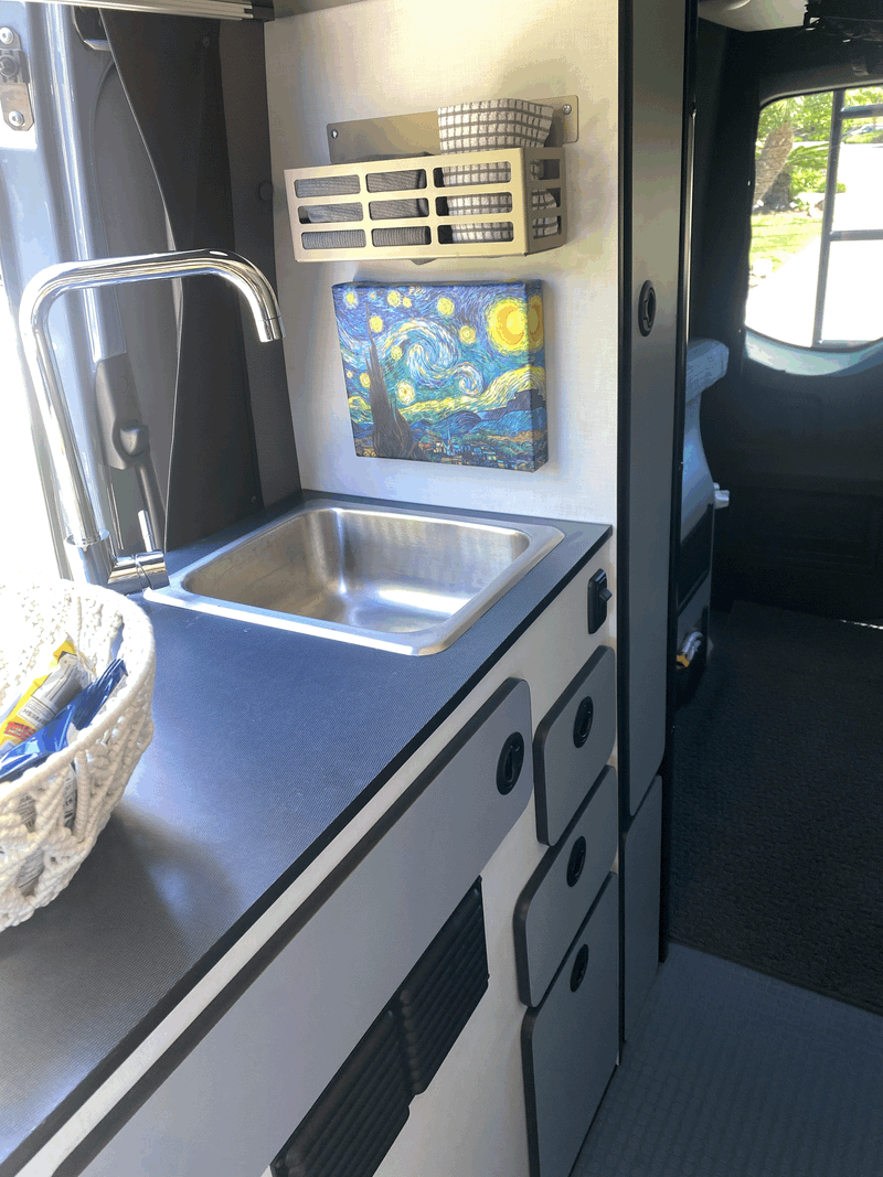 RV rental: Compact kitchenette with sink, countertop, storage drawers, and a Starry Night painting. Outside view visible. — photo 12