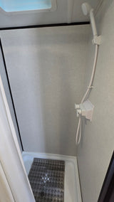 Inside an RV rental shower, showing a compact stall with light gray walls, a handheld sprayer, and a non-slip mat.