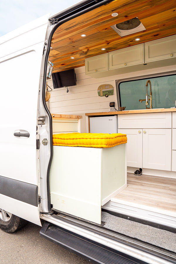 Inside a custom RV rental camper van: wooden ceiling, kitchenette, TV, and a yellow-cushioned bench visible via the open door.