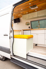 Inside a custom RV rental camper van: wooden ceiling, kitchenette, TV, and a yellow-cushioned bench visible via the open door.