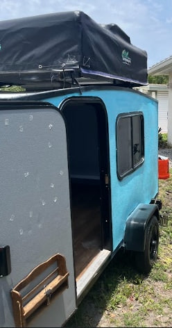 Light blue teardrop camper with rooftop tent & wooden shelf, door open. Ideal RV rental for outdoor trips!