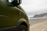 A green RV rental is parked by a scenic coastal road, overlooking the ocean and misty mountains. Perfect for road trips.