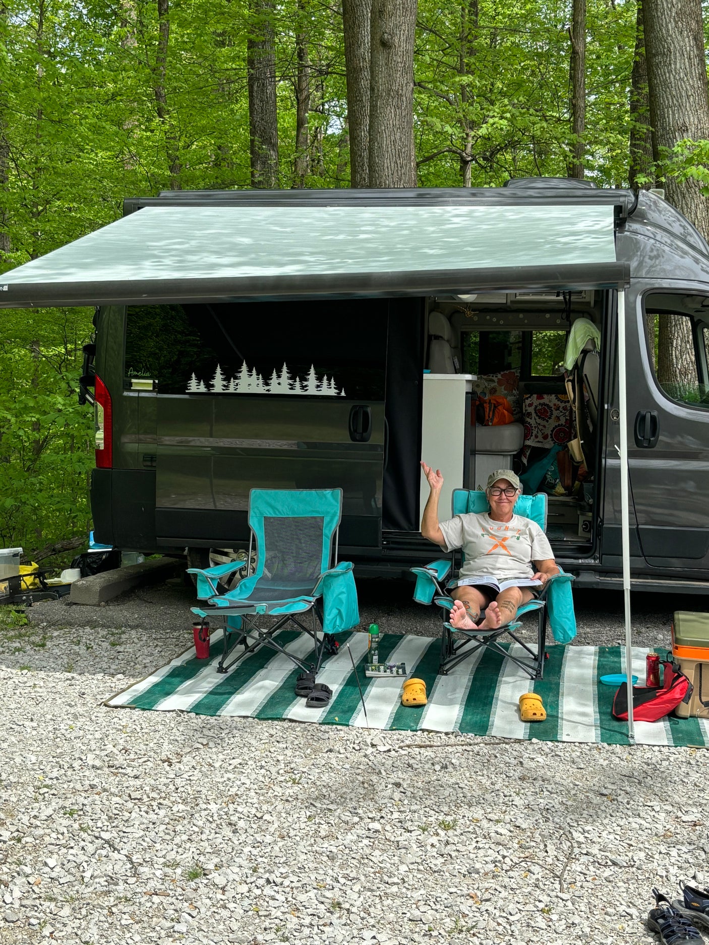 Person relaxing in a chair next to their RV rental at a peaceful wooded campsite with awning extended.