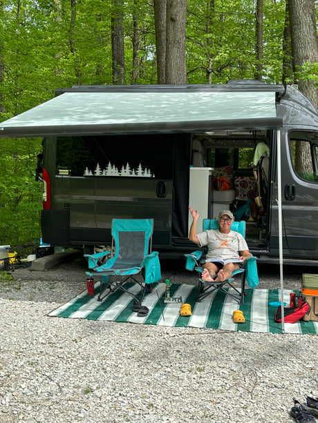 Person relaxing in a chair next to their RV rental at a peaceful wooded campsite with awning extended.