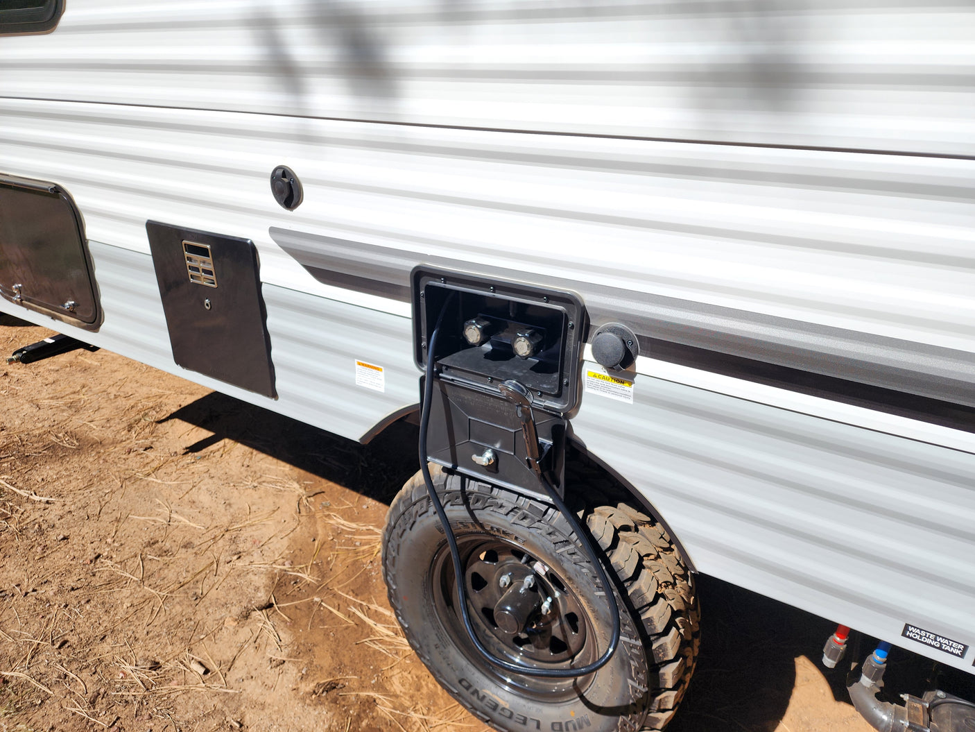 RV rental side reveals outdoor shower, fresh water fill, and waste tank hookups. Rugged tire on dirt ground.