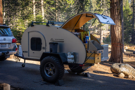 An off-road teardrop camping trailer (like an RV rental) with an open outdoor kitchen, parked with a Lexus SUV in nature.