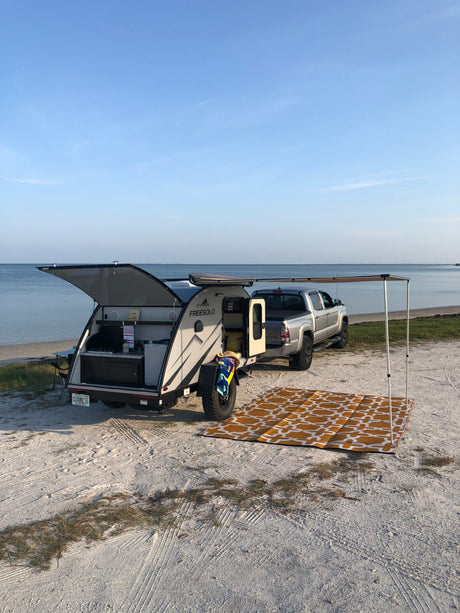 Beach camping setup with an RV rental & truck. Open camper kitchen, mat on sand by the ocean under clear skies.