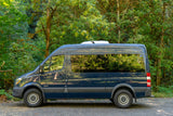 Dark blue Mercedes Sprinter RV rental parked in a lush green forest, ready for outdoor adventure.
