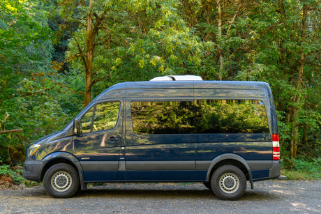 Dark blue Mercedes Sprinter RV rental parked in a lush green forest, ready for outdoor adventure.