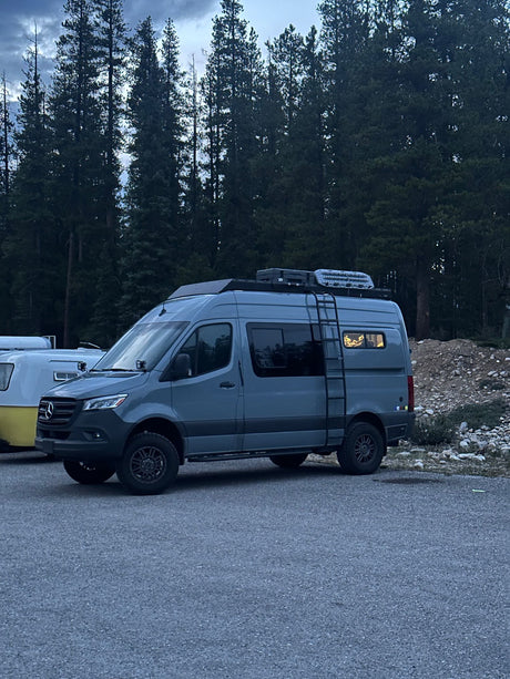 Modern gray camper van (possible RV rental) parked among tall dark trees, with a cozy light in the back window. Another camper is nearby.