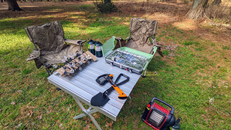 Essential camping gear for your RV rental trip: Chairs, table, stove, heater, and tools set up on grass at a campsite.