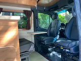 RV rental interior: Front seats and a pull-out table in a well-lit camper van with wood accents and a window view of greenery.
