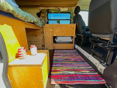 Inside an rv rental: Cozy campervan interior featuring a raised bed, wooden kitchen unit with Dometic fridge, and vibrant rag rug.