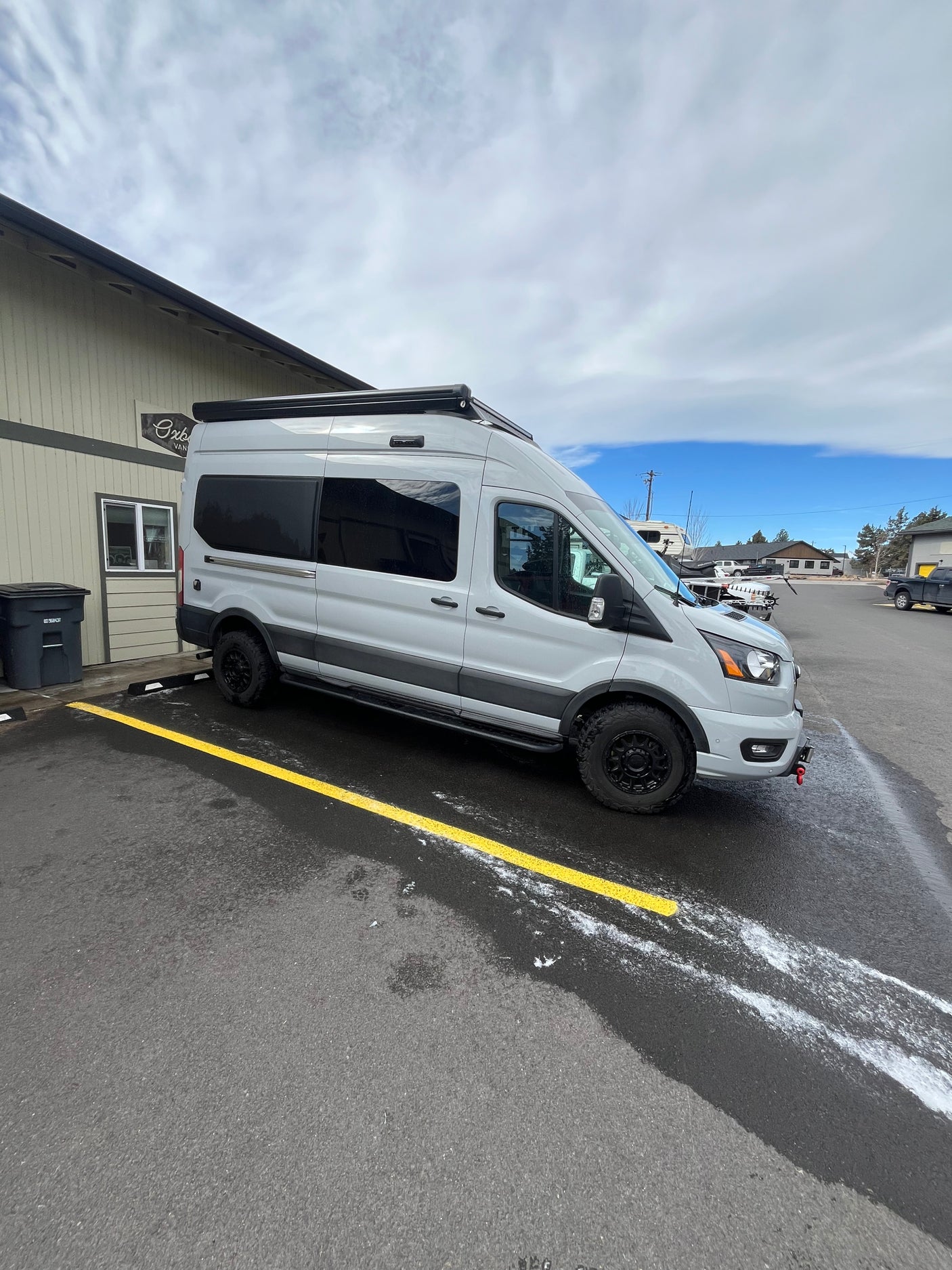 Light gray Ford Transit van (RV rental) with black wheels & roof awning, parked on an asphalt lot.