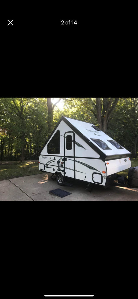 Experience nature with this A-frame pop-up camper rv rental, parked on a concrete pad amidst lush trees on a sunny day.