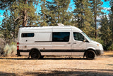 Explore nature with this white camper van RV rental, parked amidst tall pine trees on a sunny day.