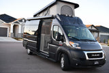 This dark grey pop-top Ram ProMaster camper van RV rental with awning & open side door is parked by a house.
