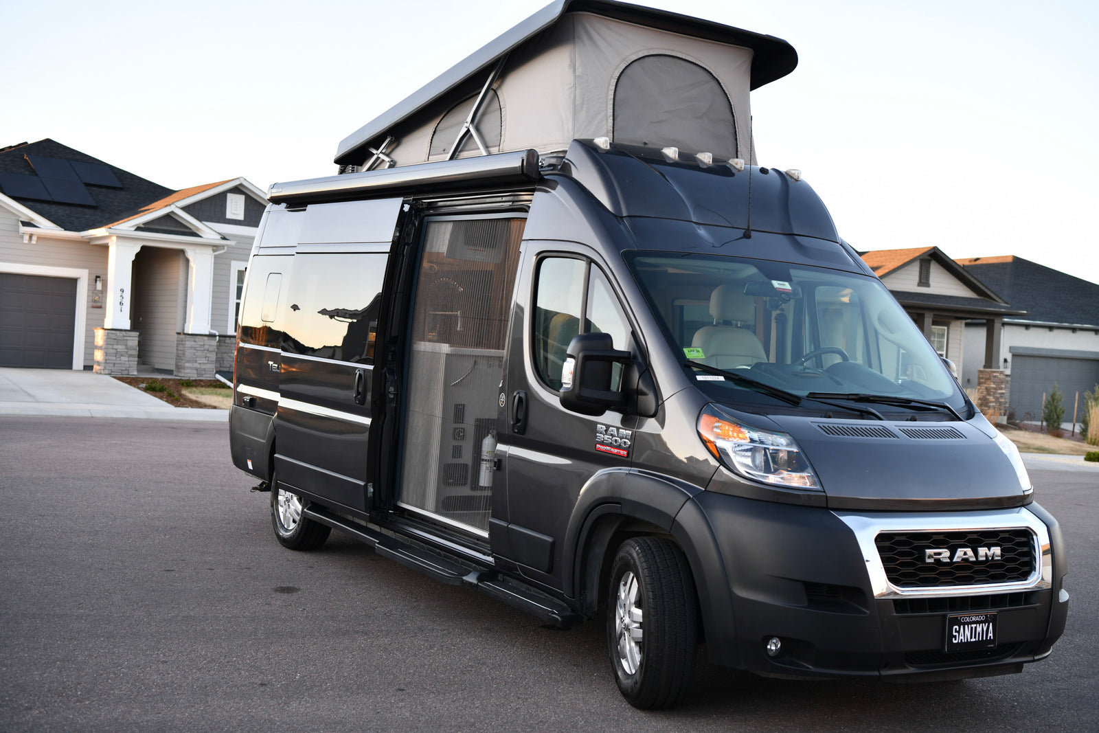 This dark grey pop-top Ram ProMaster camper van RV rental with awning & open side door is parked by a house.