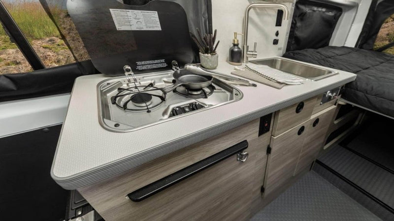 RV rental: A compact kitchen area featuring a two-burner stove, sink, and counter. A cozy bed is visible nearby. — photo 7