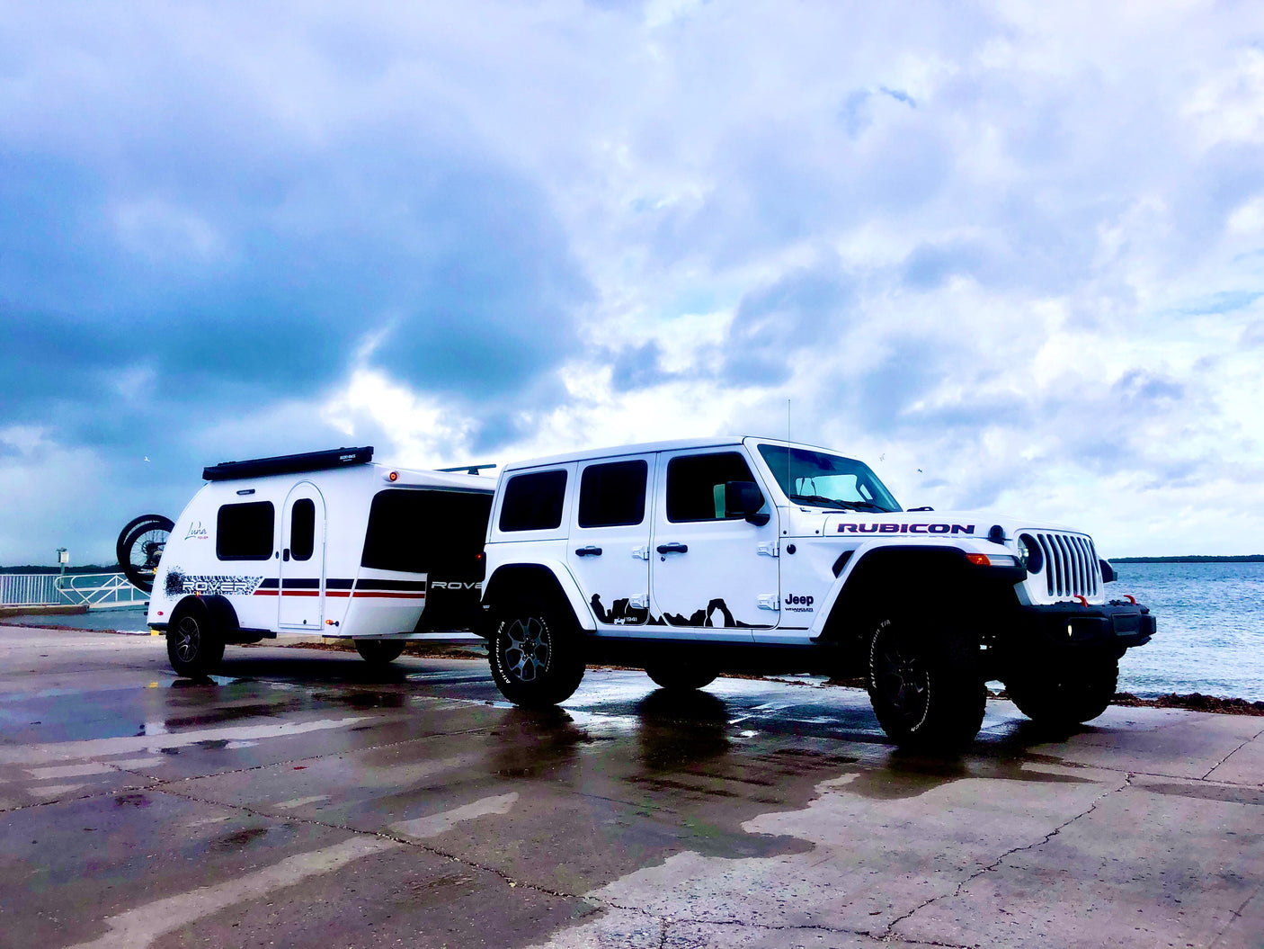 White Jeep Rubicon tows a Luna Rover teardrop RV by the water. Explore the outdoors with this ideal RV rental setup.