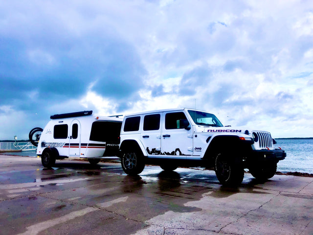 White Jeep Rubicon tows a Luna Rover teardrop RV by the water. Explore the outdoors with this ideal RV rental setup.