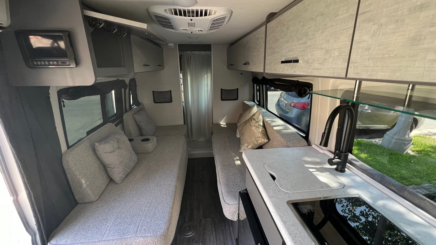 Inside a modern RV rental: light gray seating, compact kitchen with sink, overhead cabinets, and TV create a cozy living space.