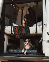 This cozy rv rental is pet-friendly! Woman reads in the upper bunk, dog relaxes on its bed in the van's open back.