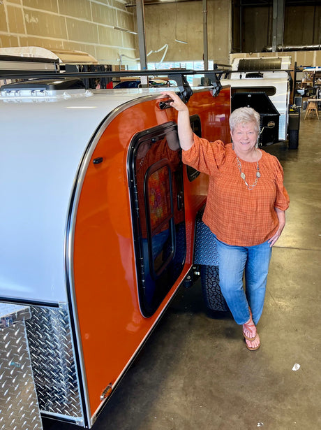 A smiling woman showcases a vibrant orange & white teardrop trailer, ideal for rv rental adventures. See this unique camper!