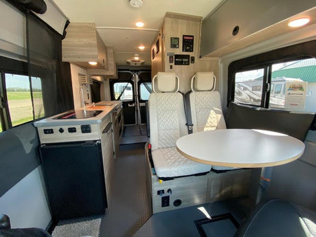 Modern RV rental interior: light wood & grey finishes, compact kitchen (sink, stove, fridge), dining table, and seats.