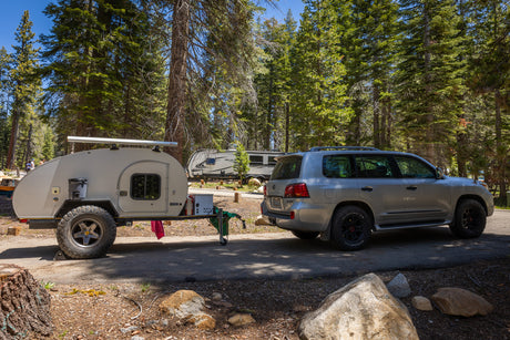 A silver SUV towing a beige teardrop trailer at a sunny forest campsite, perfect for an RV rental trip.