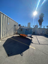 Sunny view of a blue and orange compact rv rental parked in an asphalt lot next to a metal building, with sun glare.
