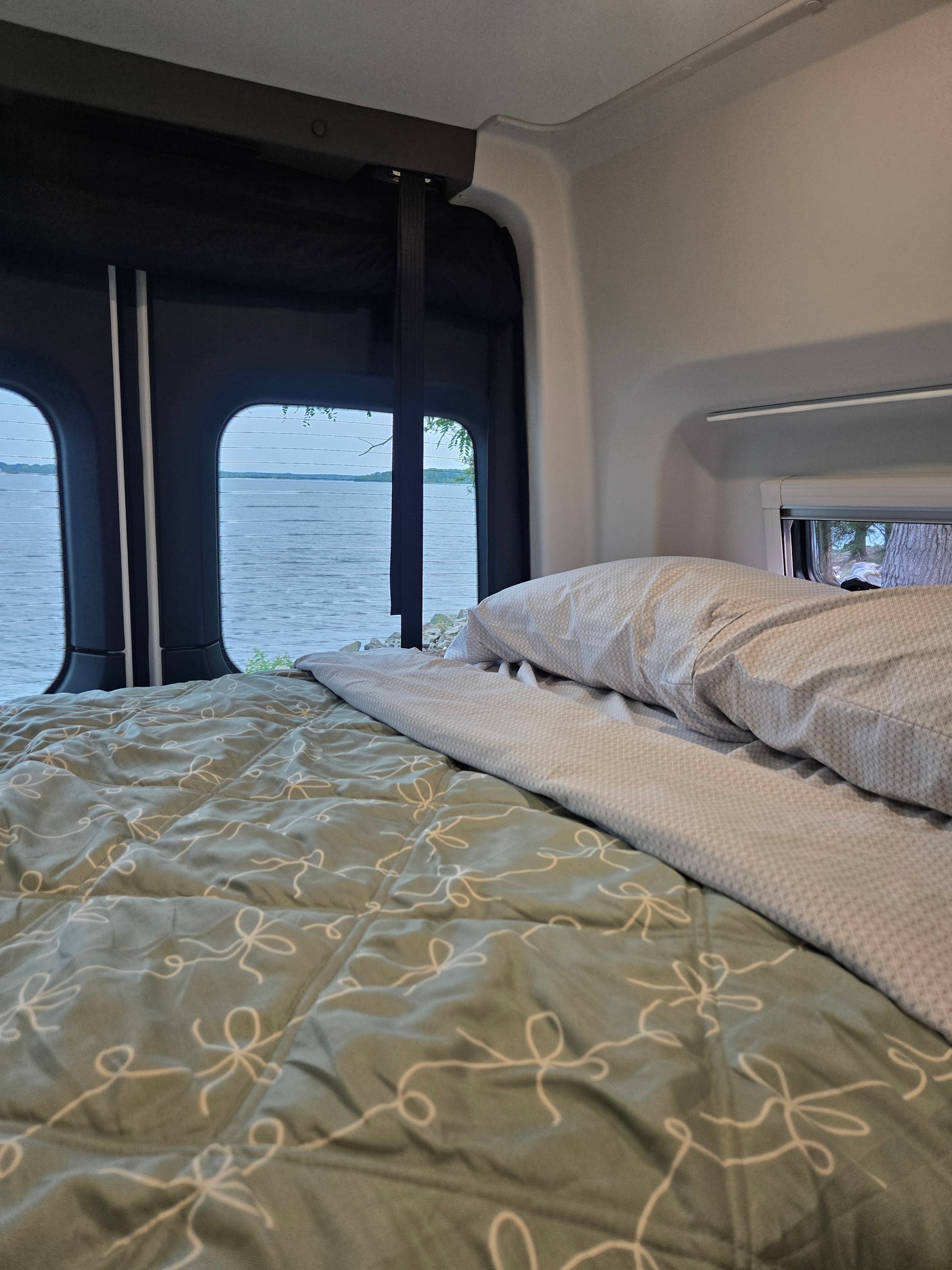 RV rental: A cozy bed with a green comforter and patterned pillow overlooks a calm lake through wide rear windows.