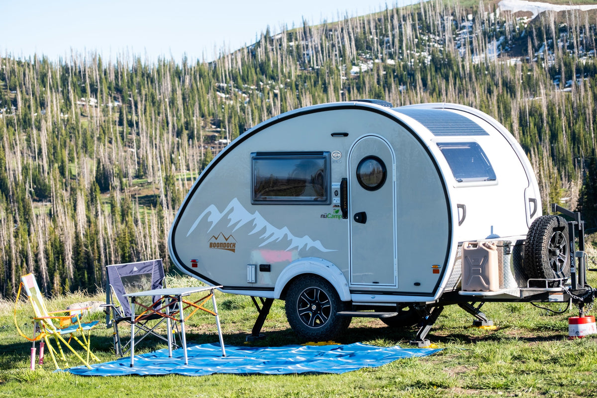 This nuCamp Boondock teardrop rv rental is set up for camping, with solar panels, chairs, and a table in a mountain forest.