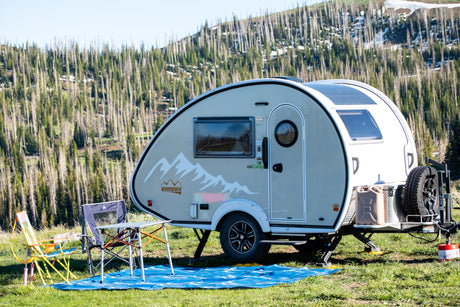 This nuCamp Boondock teardrop rv rental is set up for camping, with solar panels, chairs, and a table in a mountain forest.