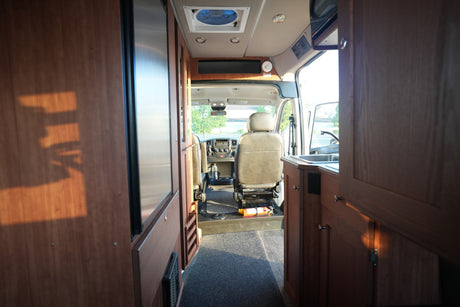 Interior of an RV rental: looking towards the cockpit, featuring wood cabinetry, a compact kitchen, and driver's seats.