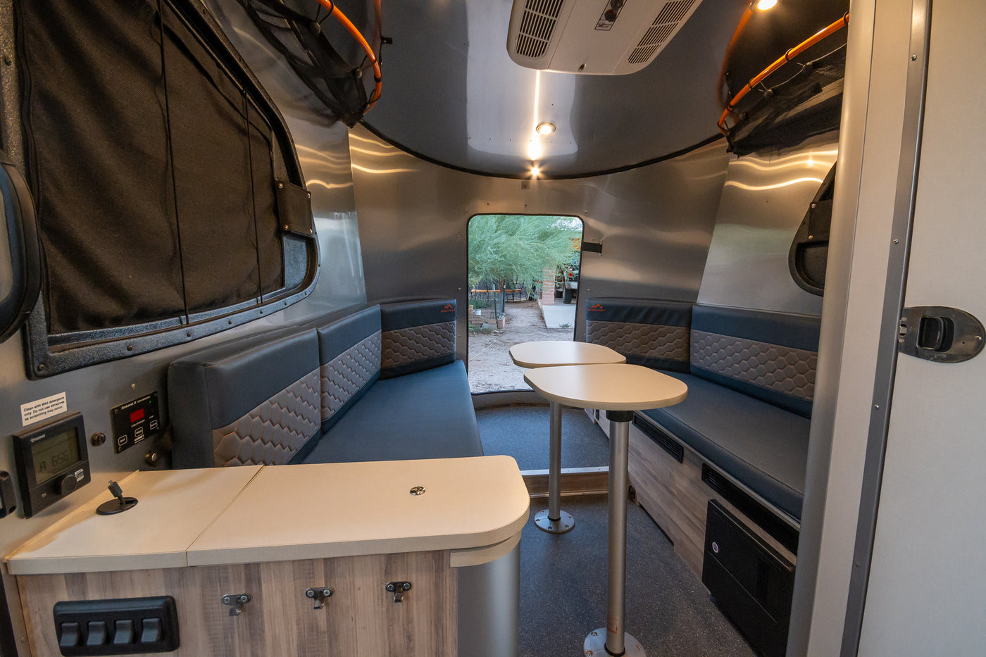 Modern RV rental interior with dinette, patterned benches, modular tables, and control panel. View outside through rear window. — photo 18
